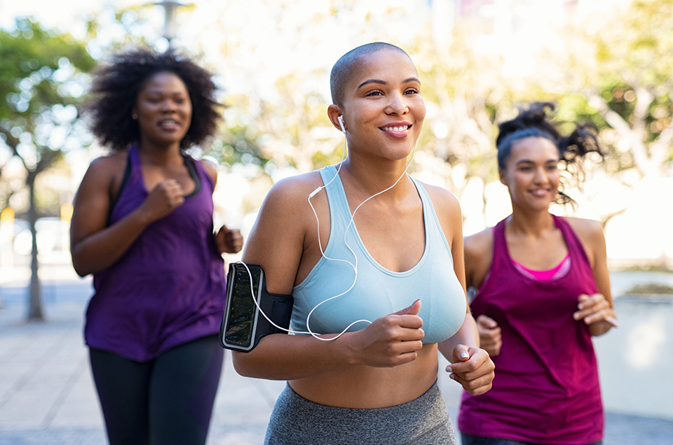 Group of curvy women jogging on track in park. Girls friends running together outdoor. Portrait of young bald woman jogging with oversize friends while listening to music on mobile phone.