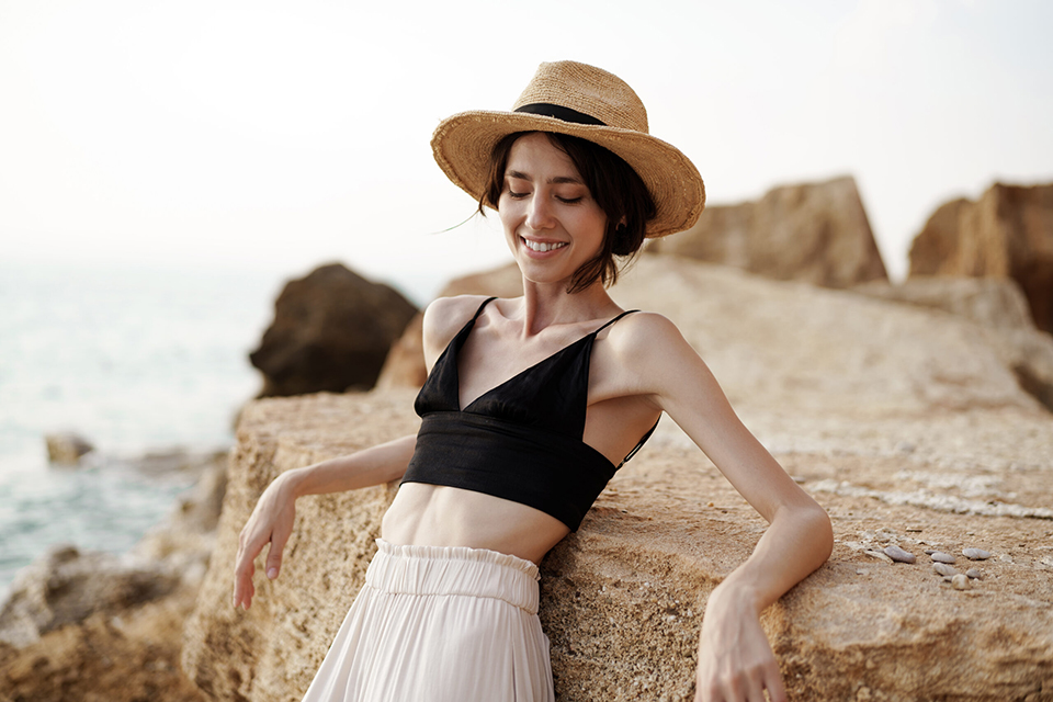 Fashion Portrait of woman in black bralette and white trousers leaning on rock at beach