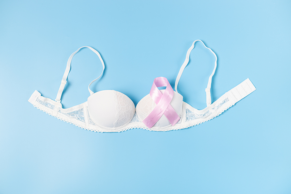 Pink ribbon emblem and white women's bra lie in the center on a blue background, close-up side view. Concept for world cancer day, breast cancer.